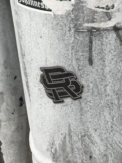 The sticker is a stylized monogram, predominantly black with a distinct metallic silver or light grey outline, giving it a slightly raised, almost three-dimensional appearance. It features two intertwined capital letters, 'P' and 'R', with the 'P' appearing to be overlaid on the 'R'. The design has a modern, somewhat gothic or ornate feel, with clean, sharp edges. It is affixed to a light grey, textured, possibly metallic pole or column, which shows signs of wear and other faint markings.