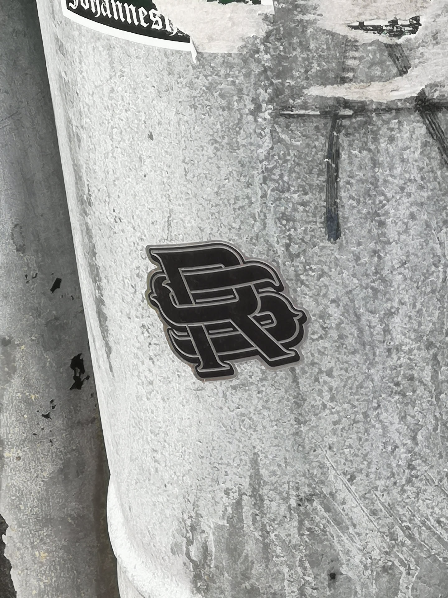 The sticker is a stylized monogram, predominantly black with a distinct metallic silver or light grey outline, giving it a slightly raised, almost three-dimensional appearance. It features two intertwined capital letters, 'P' and 'R', with the 'P' appearing to be overlaid on the 'R'. The design has a modern, somewhat gothic or ornate feel, with clean, sharp edges. It is affixed to a light grey, textured, possibly metallic pole or column, which shows signs of wear and other faint markings.