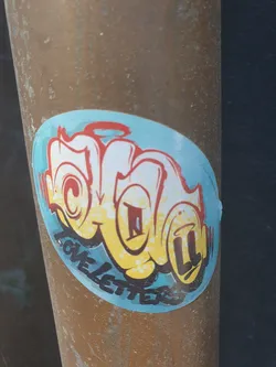 A round sticker with a graffiti-style design. The main design element appears to be stylized lettering that resembles the word "LoveLetters". The letters are in yellow and white with a blue background. There is a small halo above the design. The sticker is attached to a brown metal pole.