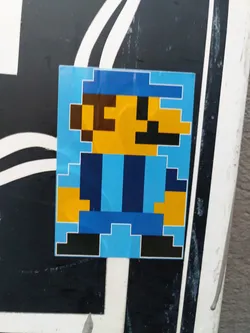 Street sticker A sticker featuring a pixelated character resembling a video game character, predominantly in blue, yellow, and black. The character has a simple design with blocky features.