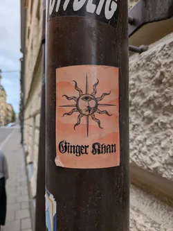 Street sticker A rectangular peach sticker on a pole. It features a black woodcut-style illustration of a sun with a human face and a third eye. Below the illustration, the text 'Ginger Khan' is written in a black gothic typeface.