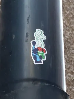Street sticker A sticker featuring pixel art of a character that seems to be boxing. The character is predominantly blue and green, with hints of red. It's affixed to a dark gray cylindrical surface, possibly a pipe or pole.