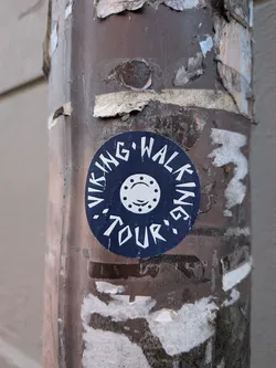Street sticker A circular sticker featuring a dark blue background with white text and a central emblem. The text 'VIKING WALKING TOUR' is arranged in an arc around the top and bottom edges of the circle. In the center, there is a white stylized circular design, possibly representing a shield boss or a wheel, with a smaller central circle and eight dots evenly spaced around it, along with two small curved lines resembling a smile or a stylized face. The sticker has a slightly worn appearance and is affixed to a heavily weathered brown pole.