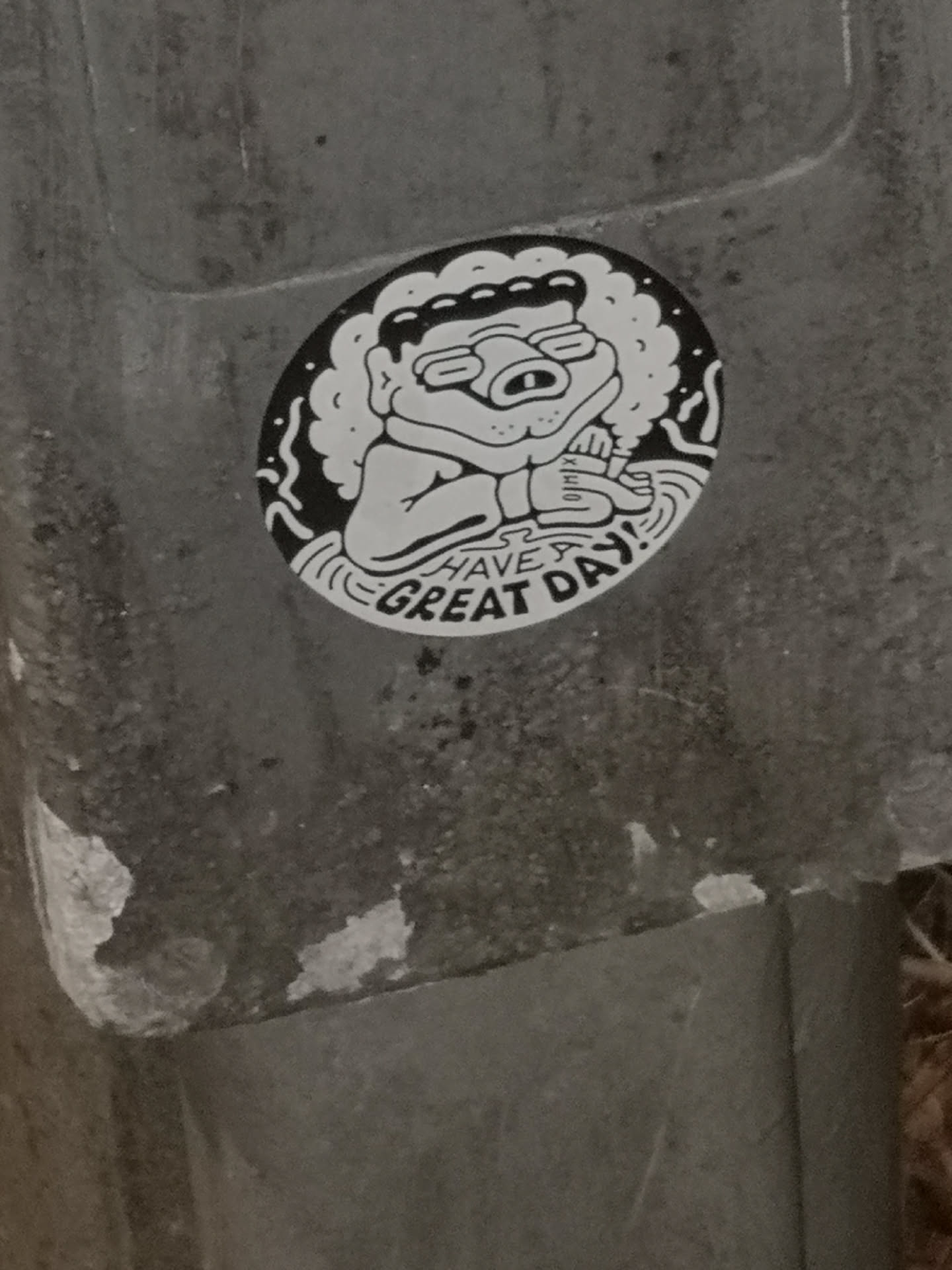 An oval-shaped sticker featuring a black and white illustration. The central figure is a cartoon character with a large head, prominent curly or afro-like hair, and thick-rimmed glasses. It has a somewhat grumpy or tired expression with a snout-like nose and a small mouth showing a single tooth. The character appears to be sitting or floating amidst wavy lines, possibly representing water or clouds. It holds a small, indistinct object in its hands. Below the character, in a bold, stylized font, is the text 'HAVE A GREAT DAY!'. The overall style is reminiscent of bold line art or a whimsical doodle.