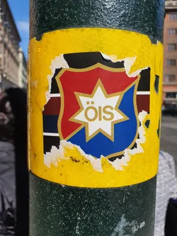 A partially torn sticker featuring the logo of ÖIS (likely a sports team or organization) on a yellow background. The logo is a shield with red, blue, and black sections, and a white star with the letters "ÖIS" in the center.