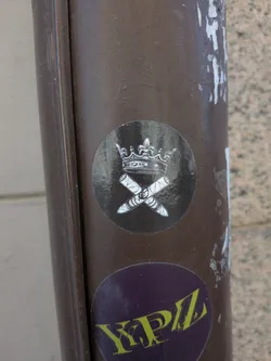 Street sticker The sticker is circular with a dark, almost black, background. In the upper half, a detailed illustration of a crown is depicted in white and light grey tones, giving it a metallic or silver appearance. Below the crown, two cigars are shown crossed over each other in an 'X' formation. These cigars are also rendered in white and grey, with visible texture and banding. A clear dollar sign symbol is visible on the band of the cigar on the right. The overall style is illustrative and somewhat ornate, creating a regal yet opulent theme.
