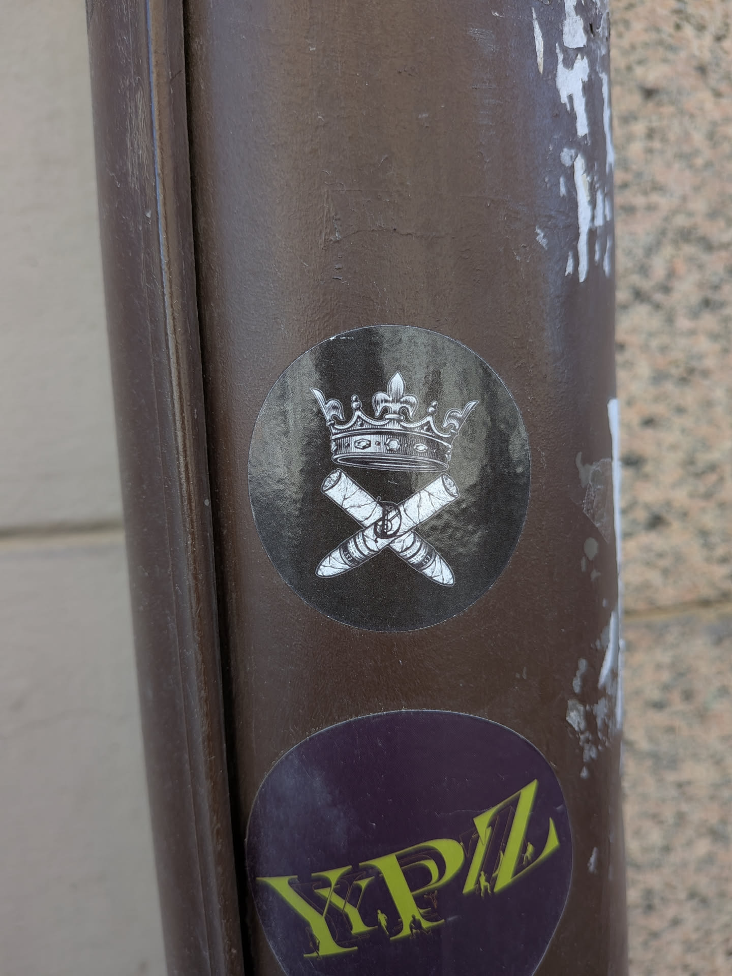 The sticker is circular with a dark, almost black, background. In the upper half, a detailed illustration of a crown is depicted in white and light grey tones, giving it a metallic or silver appearance. Below the crown, two cigars are shown crossed over each other in an 'X' formation. These cigars are also rendered in white and grey, with visible texture and banding. A clear dollar sign symbol is visible on the band of the cigar on the right. The overall style is illustrative and somewhat ornate, creating a regal yet opulent theme.