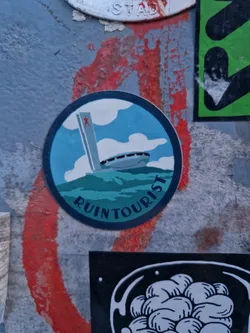 A round sticker featuring a stylized illustration of a building resembling the Palace of the Soviets, set against a teal background with a cloudy sky. The words "RUIN TOURIST" are written in a circular manner at the bottom. The sticker is adhered to a textured surface with other stickers around it.