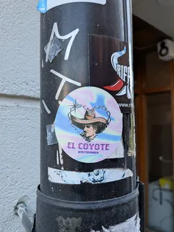 Street sticker The sticker is circular with a vibrant, iridescent, holographic background that shifts through pastel shades of pink, purple, blue, and yellow. At its center is a stylized portrait of a man, depicted from the chest up, looking slightly to the right. He wears a wide-brimmed, dark brown sombrero with intricate orange and black detailing on the brim. A thin, dark chain forms a heart shape around his head, connecting to a stylized four-pointed star at the top. The man has a dark mustache and a serious, somewhat enigmatic expression. Below the portrait, in a blocky, pixelated purple font, is the text "EL COYOTE", and beneath that, in a smaller, similar font, "@VESTAYINDEN". The overall aesthetic blends traditional imagery with a modern, vaporwave-like style.