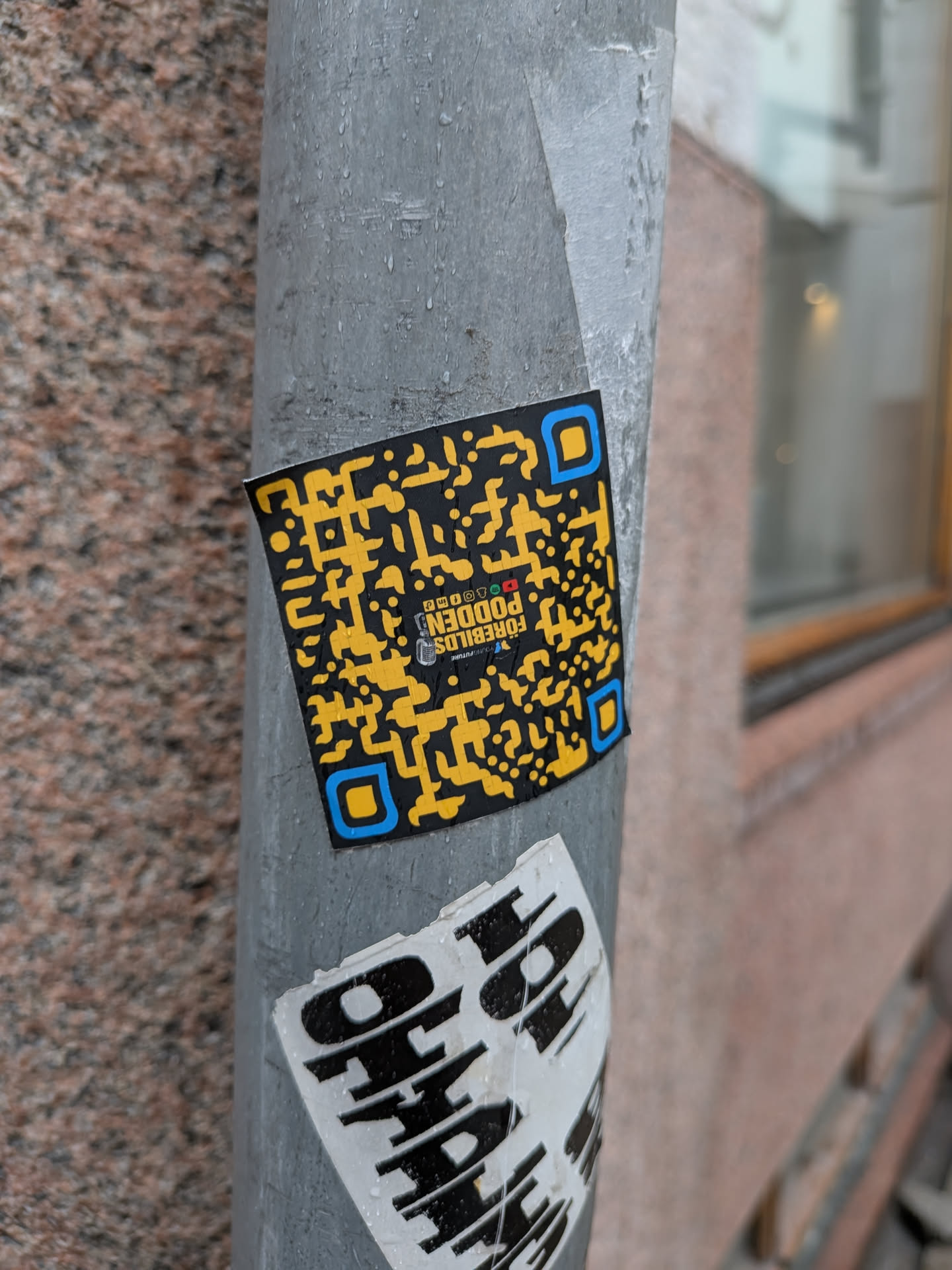 A square-shaped sticker featuring a highly stylized QR code. The primary colors are black for the background and a bright yellow for the QR code modules. The three corner finder patterns of the QR code are distinctly outlined in a bright blue, with their inner squares remaining yellow. Embedded within the center of the QR code pattern, oriented upside down, is the text 'FÖREBILDS PODDEN'. Below this, smaller text reads 'YOUR FUTURE' and 'PODDEN.FI'. There are also tiny words 'PHOTO' and 'BLOGG' visible. The sticker is slightly wet with visible water droplets and is affixed to a textured grey metal pole.