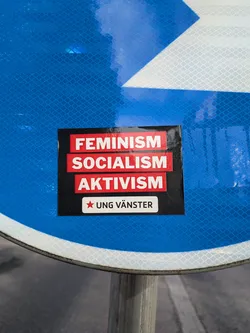 Street sticker A rectangular black sticker featuring three red horizontal blocks with the white text 'FEMINISM', 'SOCIALISM', and 'AKTIVISM'. Below these, a white block contains a red five-pointed star and the text 'UNG VÄNSTER'.