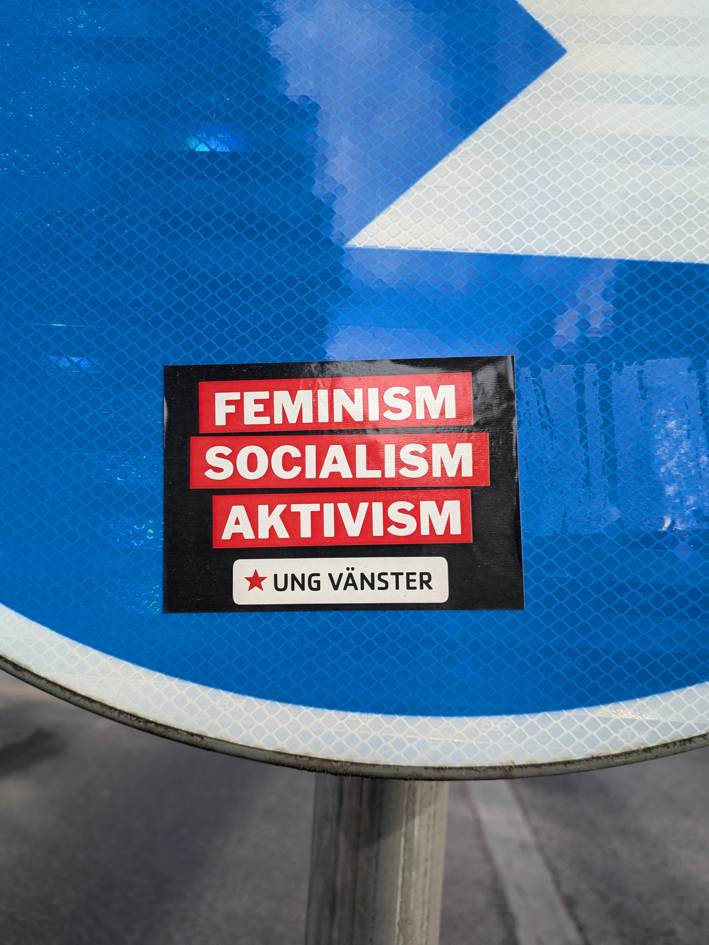A rectangular black sticker featuring three red horizontal blocks with the white text 'FEMINISM', 'SOCIALISM', and 'AKTIVISM'. Below these, a white block contains a red five-pointed star and the text 'UNG VÄNSTER'.