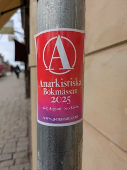 Street sticker The sticker is on a gray pole. It has a red top and a pink/purple bottom with white text. The symbol for Anarchy is at the top.