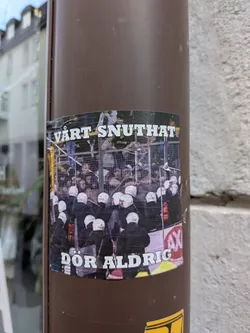 A rectangular sticker depicting a crowd of riot police wearing white helmets and dark uniforms, viewed from behind near a metal fence. Bold white text with black outlines is positioned at the top and bottom.