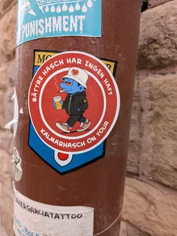 A circular sticker with a red background and a white border. It depicts a blue Smurf character with bloodshot eyes, smoking a joint and holding a cup of beer. The character is dressed in casual/ultras style, wearing a white bucket hat with a Kalmar FF crest, a black hoodie with a Stone Island-style compass patch on the sleeve, black pants, and red sneakers. White Swedish text arches around the inner border.