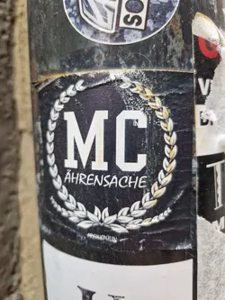 Street sticker Black and white sticker with the letters MC in the center, surrounded by a laurel wreath. The text Ährensache is written below the letters. There is a small text at the bottom that seems to be a signature or a watermark.