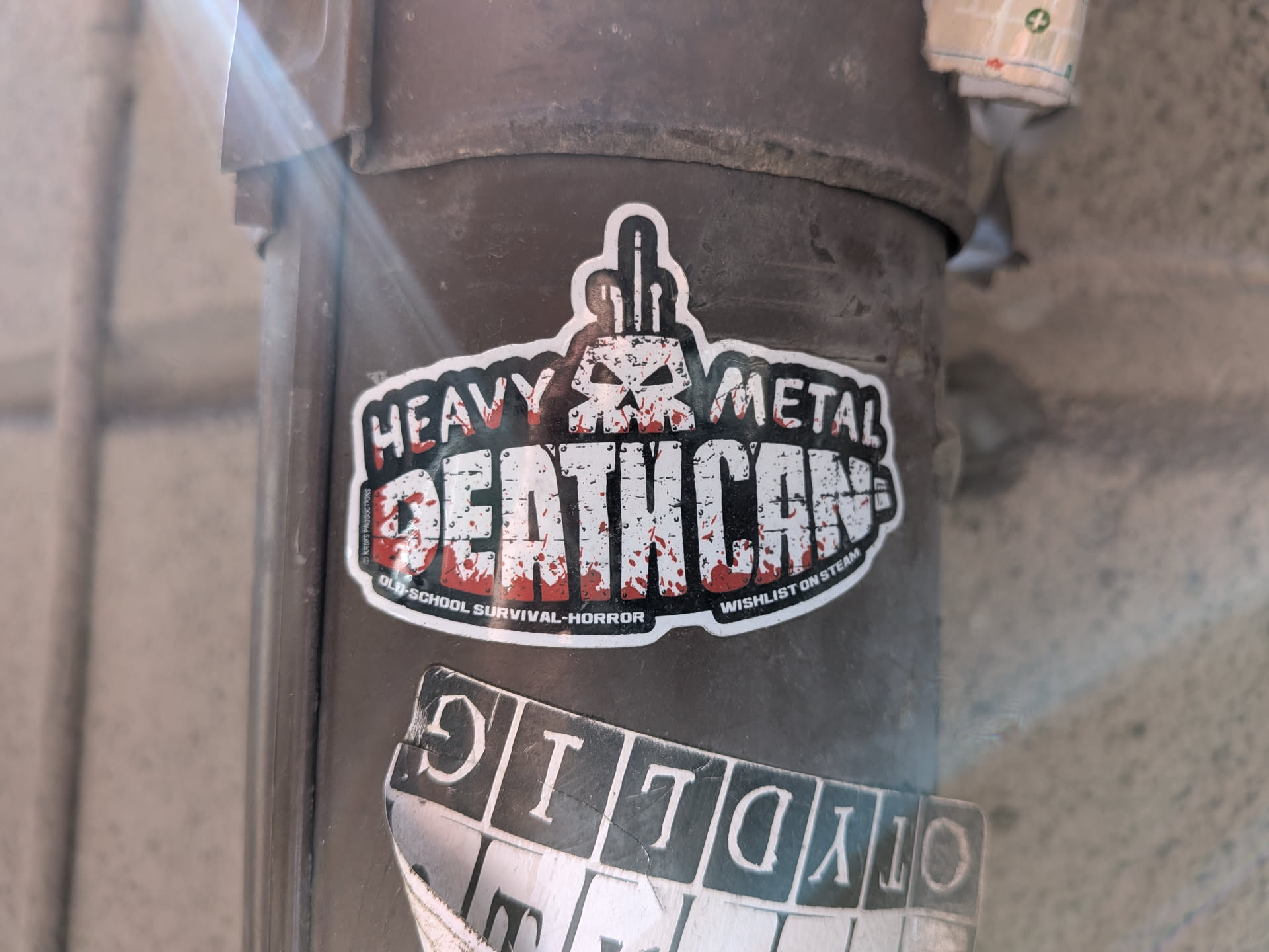 A die-cut sticker featuring a stylized skull with a middle-finger gesture on top. The main text reads 'HEAVY METAL DEATH CAN' in a distressed, blood-splattered font. Subtext promotes it as an 'old-school survival-horror' game to wishlist on Steam.