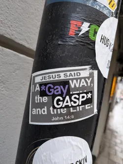 Street sticker The central sticker is a rectangular design with a white border, featuring a dark grey or black background. At the top, 'JESUS SAID' is printed in white capital letters on a lighter grey bar. Below this, the main text reads 'I A*Gay WAY,' with the word 'Gay' highlighted in a vibrant purple. Following this, 'the GASP*' is displayed prominently, with 'GASP' in large white capital letters on a black rectangular patch. The phrase concludes with 'and the Life.' At the bottom, 'John 14:6' is written in smaller white text, referencing the original scripture. The sticker shows signs of wear, with some minor wrinkles and slight peeling, indicating it is a physical sticker adhered to a surface.