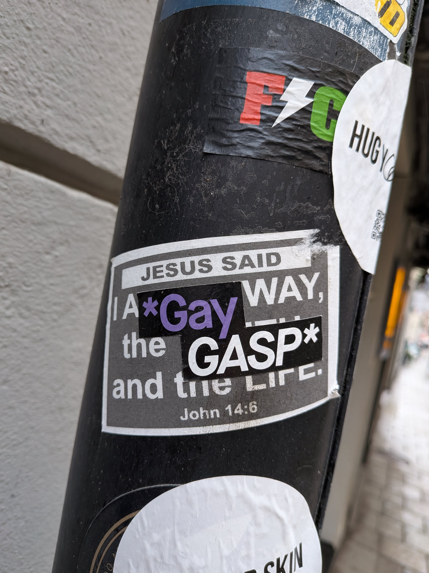 The central sticker is a rectangular design with a white border, featuring a dark grey or black background. At the top, 'JESUS SAID' is printed in white capital letters on a lighter grey bar. Below this, the main text reads 'I A*Gay WAY,' with the word 'Gay' highlighted in a vibrant purple. Following this, 'the GASP*' is displayed prominently, with 'GASP' in large white capital letters on a black rectangular patch. The phrase concludes with 'and the Life.' At the bottom, 'John 14:6' is written in smaller white text, referencing the original scripture. The sticker shows signs of wear, with some minor wrinkles and slight peeling, indicating it is a physical sticker adhered to a surface.