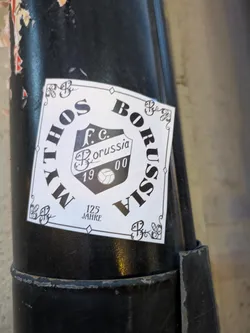 Street sticker A white, rectangular sticker with a black border. In the center is a shield-like emblem in black and white, containing the text 'F.c. Borussia', the year '1900', and a stylized soccer ball. The words 'MYTHOS' are curved along the top left of the sticker and 'BORUSSIA' along the top right, with '125 JAHRE' (125 Years) below the emblem. Decorative flourishes are present in the corners and around the text.