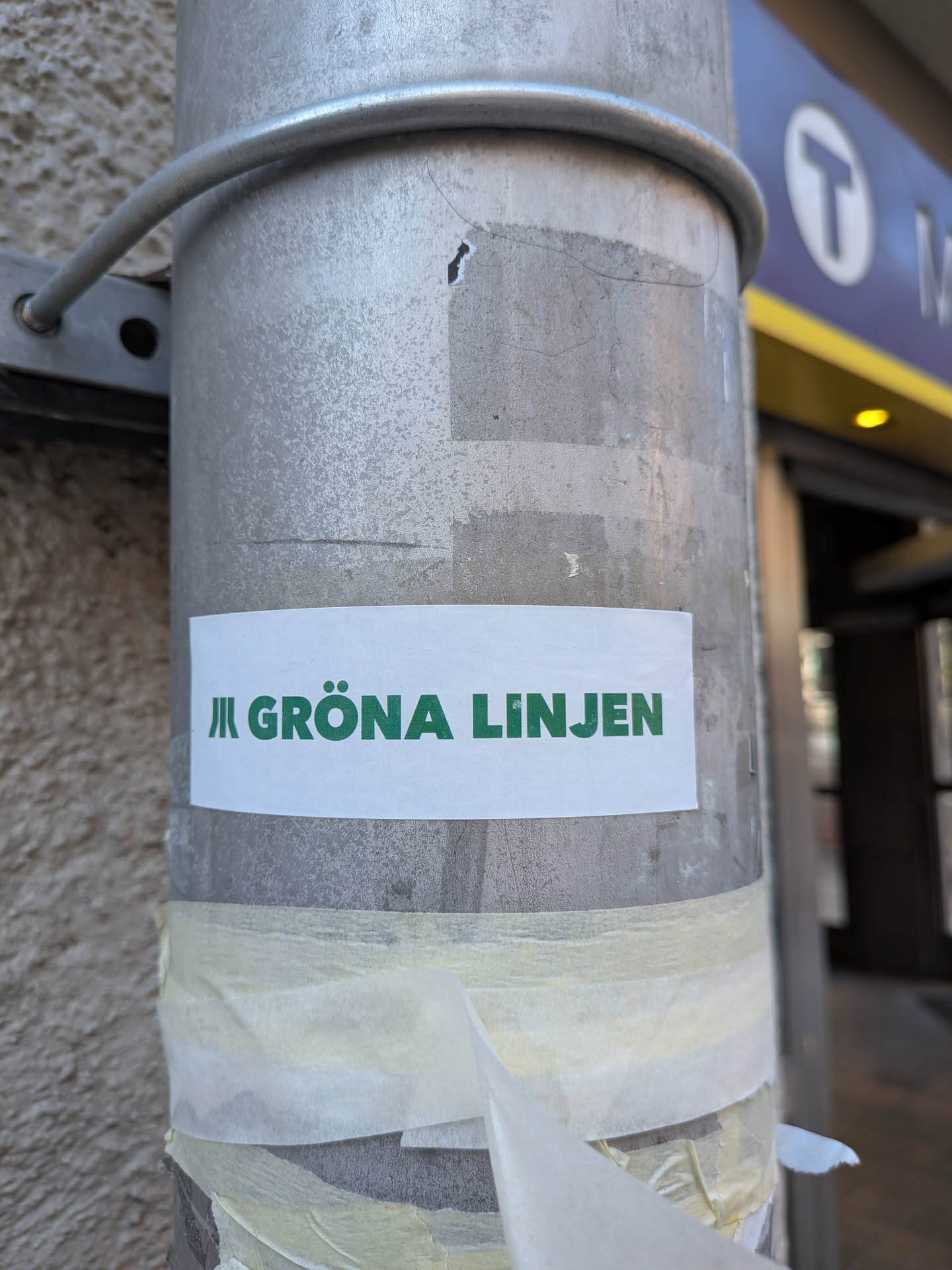 The sticker is a rectangular white label featuring bold, uppercase green text that reads 'GRÖNA LINJEN'. To the left of the text, there is a stylized symbol composed of three vertical green lines of increasing height, commonly associated with metro or subway systems. The sticker is applied horizontally to a textured, metallic grey pole, with remnants of old, torn masking tape visible beneath it. The white background of the sticker provides a stark contrast to the vibrant green lettering.
