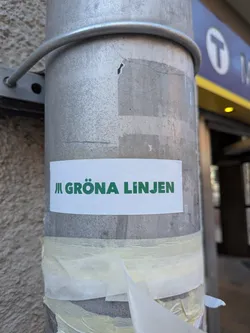 Street sticker The sticker is a rectangular white label featuring bold, uppercase green text that reads 'GRÖNA LINJEN'. To the left of the text, there is a stylized symbol composed of three vertical green lines of increasing height, commonly associated with metro or subway systems. The sticker is applied horizontally to a textured, metallic grey pole, with remnants of old, torn masking tape visible beneath it. The white background of the sticker provides a stark contrast to the vibrant green lettering.
