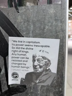Street sticker The sticker is a black and white design featuring a quote from Ursula K. Le Guin about capitalism and resisting power. It also shows a portrait of the author and includes a logo and the text 'Allt åt alla'. The sticker is affixed to a textured, possibly concrete or stone, surface.