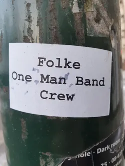 Street sticker A sticker featuring the text "Folke One Man Band Crew" in a typewriter-style font. The sticker is affixed to a dark-green pole. 