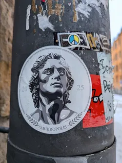 The sticker is circular, featuring a detailed grayscale illustration of a classical-style bust of a young man. He has curly hair and wears a laurel wreath on his head, looking upwards and slightly to the right. The artwork uses strong contrasts of black, white, and various shades of gray to create depth and a sculptural effect. The bust is set against a lighter gray background, and the entire design is framed by a decorative border that resembles the rim of an ancient coin or medallion. At the bottom, within this border, the word 'ABIKROPOLIS' is written, flanked by small laurel branch motifs. The numbers '20' and '25' are subtly placed on the left and right sides, respectively, near the inner edge of the circular border.