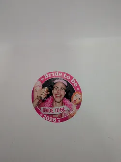 Street sticker Circular pink sticker depicting a smiling man with a pink tiara, feather boa, and 'BRIDE TO BE' sash. He is flanked by novelty party inflatables. The border features the text 'Bride to be' and '2026' with small hearts.