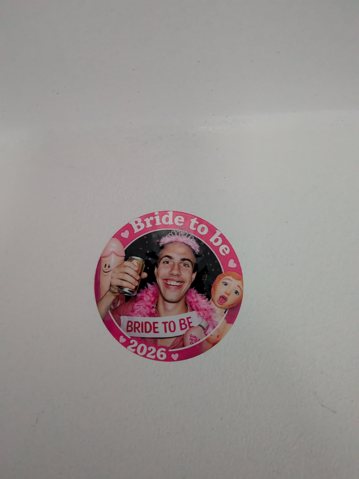 Circular pink sticker depicting a smiling man with a pink tiara, feather boa, and 'BRIDE TO BE' sash. He is flanked by novelty party inflatables. The border features the text 'Bride to be' and '2026' with small hearts.
