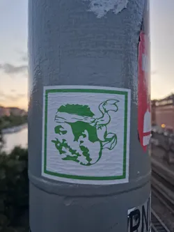 Street sticker A green creature is depicted in a minimalist style on a white background, framed by a green square. The creature appears to be a stylized figure with a headband or bandana and possibly wings or appendages. The style suggests a possible reference to street art or graphic design. The sticker is affixed to a gray pole, likely outdoors.