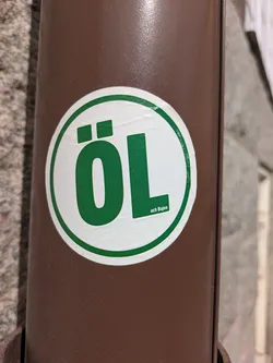 Street sticker The sticker is a white circle with a green border, displaying the word "ÖL" in large, green letters. Below "ÖL", in smaller green text, it says "och Bajen". The sticker is affixed to a brown metal drainpipe.