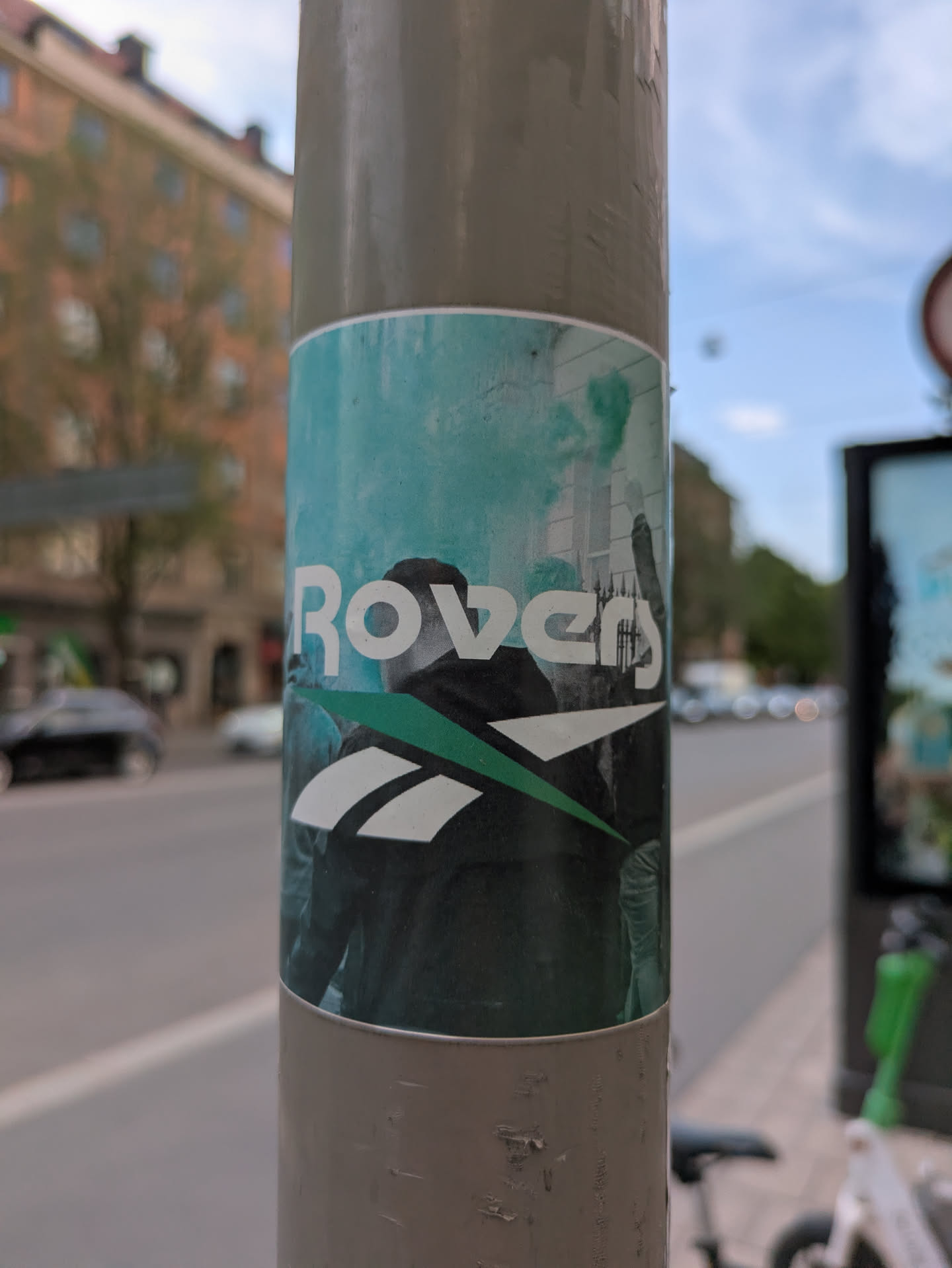 Street sticker The sticker shows the word "Rover" in white lettering, with the Reebok logo replacing the letter "o". The background features an image of a group of people, possibly at a protest or event, with green smoke or flares. The sticker is affixed to a gray metal light pole, with an urban street scene visible in the background.