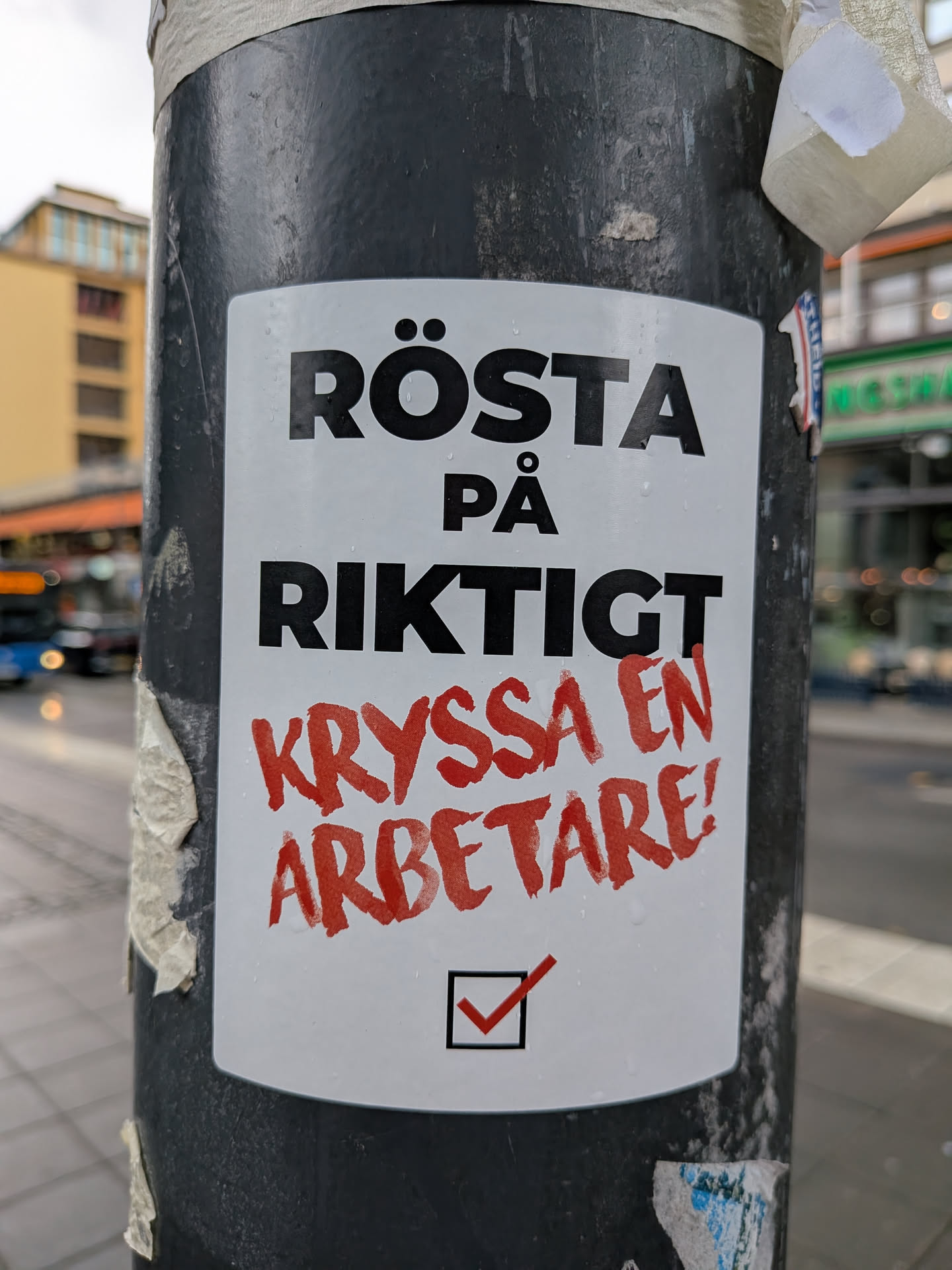 White rectangular sticker on a pole featuring black text 'RÖSTA PÅ RIKTIGT', red brush-style text 'KRYSSA EN ARBETARE!', and a checkbox with a red checkmark.