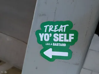 The sticker is irregularly shaped, resembling a green blob or cloud with a thin white outline. It features bold white text in a casual, slightly brush-stroke style font. The top line reads "TREAT", the middle line "YO' SELF", and a smaller, italicized line below that says "LIKE A BASTARD". Below the text, there is a prominent white arrow pointing to the left. The sticker is applied to a grey, weathered concrete or metal surface.