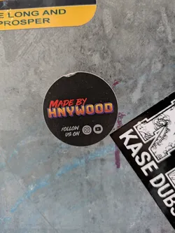 Street sticker A round black sticker with the text "MADE BY HNYWOOD" in orange and yellow.  Below the text, it says "FOLLOW US ON" with Instagram and YouTube icons.