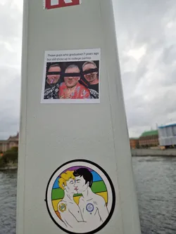 Street sticker A sticker featuring a drawing of two men kissing, set against a rainbow background.  Another sticker above it shows a photo of three men, likely at a party. 