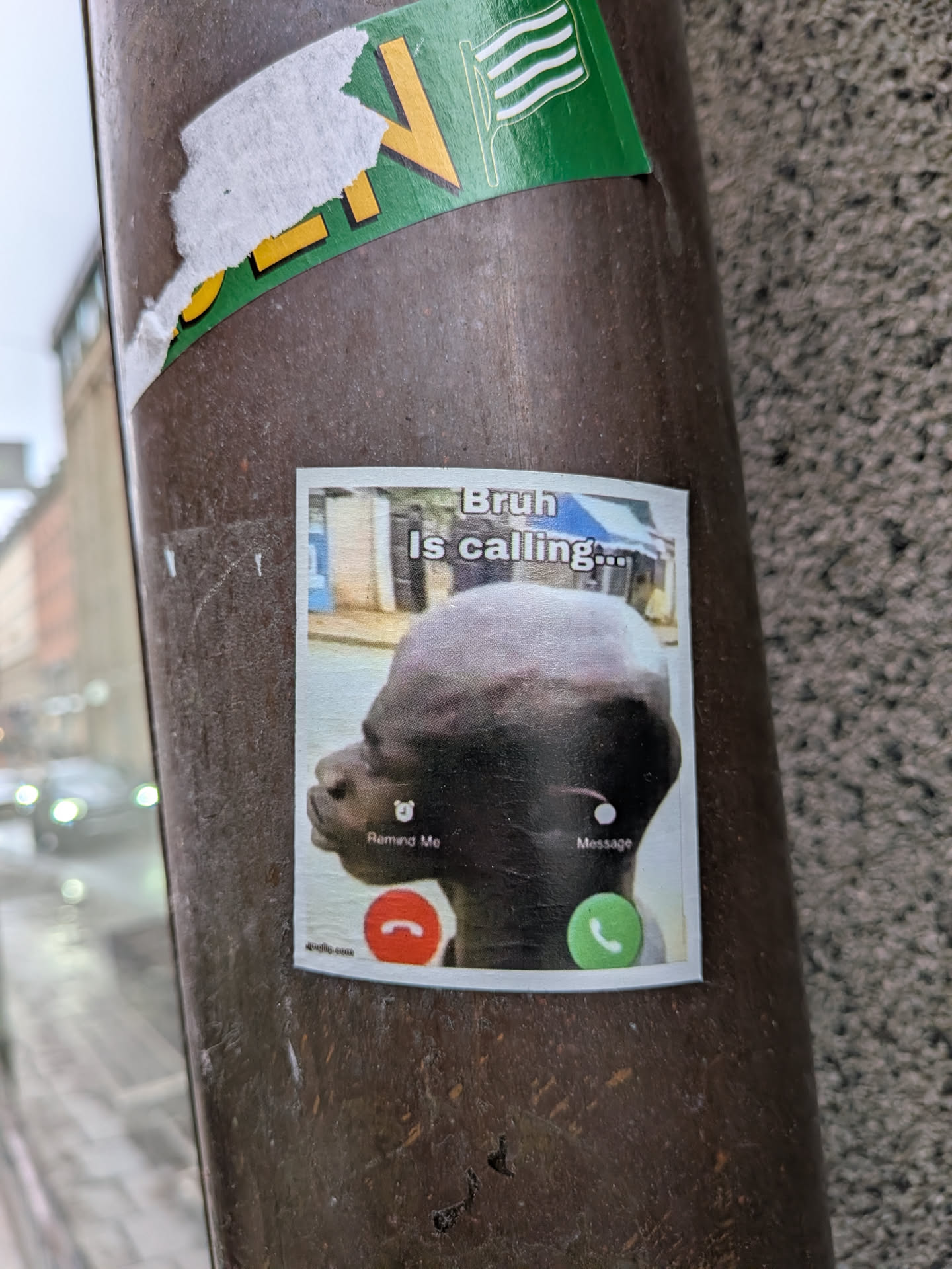A rectangular sticker simulating a smartphone's incoming call screen. At the top, in a white box, the text 'Bruh Is calling...' is displayed in black. Below this, a large, somewhat blurry image of a person's dark-skinned, bald head is shown in profile, set against a blurry background of buildings. At the bottom left, there's a small alarm clock icon with 'Remind Me' text, and at the bottom right, a small white circle with 'Message' text. Below these options are two prominent circular buttons: a red one on the left with a white phone hang-up icon, and a green one on the right with a white phone answer icon. The sticker has a white border and appears slightly worn.