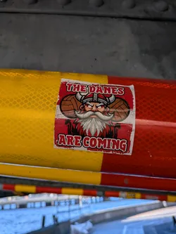 Street sticker The sticker features a stylized illustration of a Viking character with a prominent white beard and mustache, wearing a horned helmet. Behind the Viking's head are two brown, textured spheres that resemble old-fashioned basketballs. The background of the sticker is divided into horizontal stripes, with the top section featuring the Danish flag (red with a white cross) and the bottom section in a solid red color. The text 'THE DANES' is prominently displayed in a distressed, white font with a red outline above the Viking's head, and 'ARE COMING' is displayed in a similar font below the Viking. The overall aesthetic is bold and somewhat vintage.