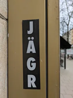 Street sticker This is a black rectangular sticker with the word "JÄGR" printed vertically in white sans-serif font. The sticker is placed on a gold surface. It could be related to the drink Jägermeister, but it's slightly different.
