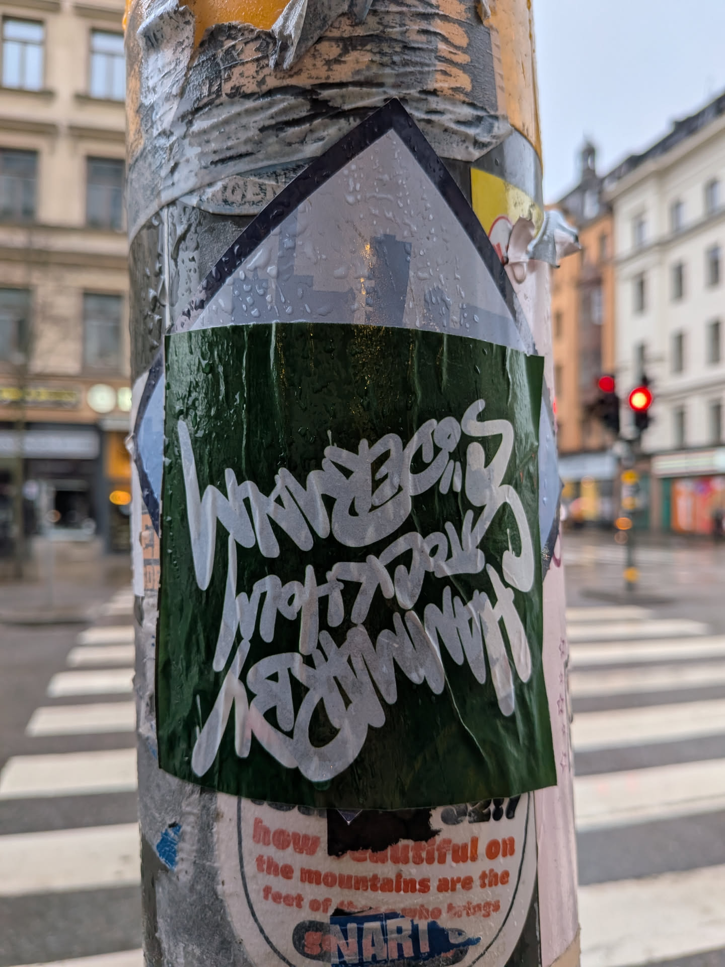 The sticker is rectangular with a pointed top, resembling a house or arrow shape, and is affixed to a metal pole covered in other worn stickers and tape. The main body of the sticker is a dark, forest green color, appearing somewhat textured and wet with numerous water droplets. Overlaid on this green background is stylized white text, written in a distinct graffiti handstyle. The text reads 'WANDERS' on the top line, 'STOCKHOLM' on the middle line, and 'GRAFFITI' on the bottom line. The white lettering has a slightly translucent quality, allowing some of the green background to show through, especially where the water droplets are. The sticker is slightly creased and shows signs of wear, indicating it has been exposed to the elements.