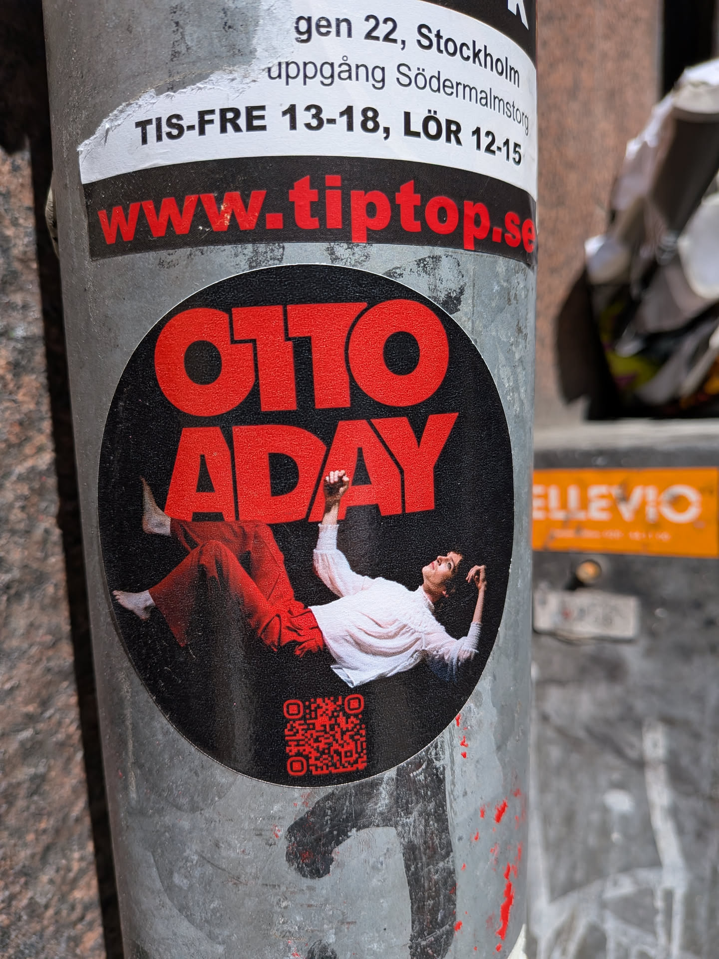 The central sticker is circular with a matte black background. At the top, the name "OTTO ADAY" is prominently displayed in large, bold, red sans-serif capital letters. Below the text, a person is depicted lying on their back, looking upwards, with their arms slightly raised. The person is wearing loose-fitting red pants and a light-colored (likely white or cream) long-sleeved shirt, with bare feet visible. In the bottom right quadrant of the black circle, there is a small, red QR code. The overall design is modern and eye-catching, using a strong contrast between red and black.