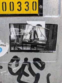 A rectangular black-and-white sticker showing an older man talking on a telephone behind a desk in an office. Features the text 'KJØBENHAVNS ÆLDRE' at the bottom right.