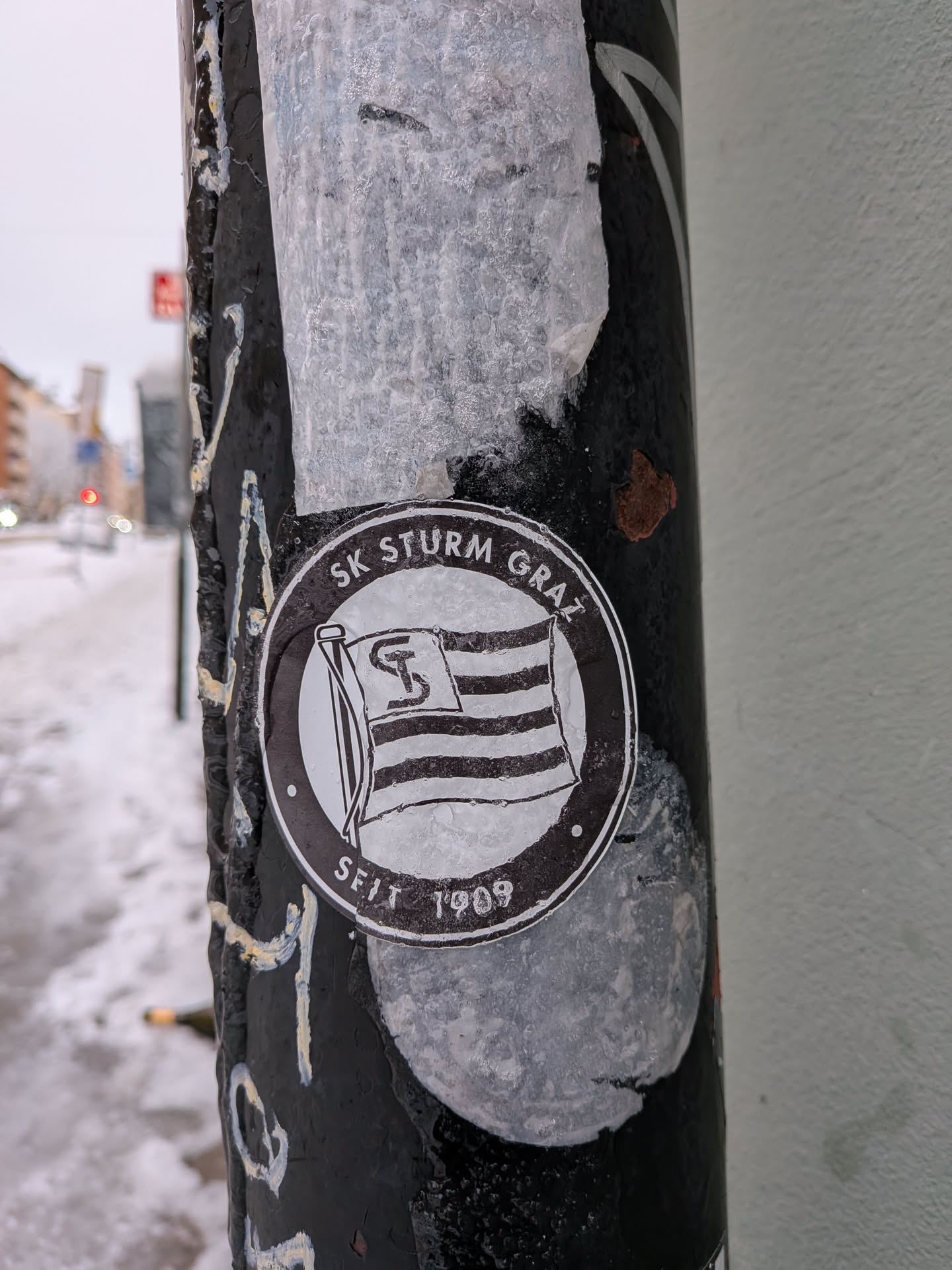 A circular, black and white sticker featuring the emblem of SK Sturm Graz. The central design depicts a waving flag with horizontal stripes, upon which a stylized 'S' and 'K' (or a unique 'S' logo) are prominently displayed. An outer ring frames the flag, with the text 'SK STURM GRAZ' arching across the top and 'SEIT 1909' (meaning 'Since 1909') at the bottom. The sticker appears slightly worn and is partially covered by a thin layer of ice or moisture, giving it a textured, frosted appearance. It is affixed to a dark, possibly metal, pole.