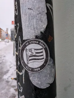 Street sticker A circular, black and white sticker featuring the emblem of SK Sturm Graz. The central design depicts a waving flag with horizontal stripes, upon which a stylized 'S' and 'K' (or a unique 'S' logo) are prominently displayed. An outer ring frames the flag, with the text 'SK STURM GRAZ' arching across the top and 'SEIT 1909' (meaning 'Since 1909') at the bottom. The sticker appears slightly worn and is partially covered by a thin layer of ice or moisture, giving it a textured, frosted appearance. It is affixed to a dark, possibly metal, pole.