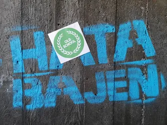 A square white sticker featuring a prominent circular design in the center. The circle is a vibrant lime green, bordered by a white laurel wreath. Inside the wreath, the words 'OLD SCHOOL' are stacked vertically in clean, white, sans-serif capital letters. The sticker is placed on a rough, dark grey concrete wall, partially obscuring blue spray-painted graffiti.