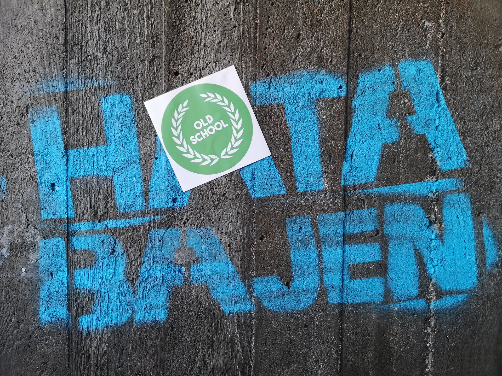 A square white sticker featuring a prominent circular design in the center. The circle is a vibrant lime green, bordered by a white laurel wreath. Inside the wreath, the words 'OLD SCHOOL' are stacked vertically in clean, white, sans-serif capital letters. The sticker is placed on a rough, dark grey concrete wall, partially obscuring blue spray-painted graffiti.