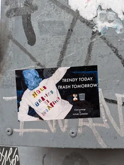 Street sticker The sticker features a central image of a person wearing a light grey long-sleeved sweatshirt, with their head obscured by a crumpled blue plastic bag. The sweatshirt has colorful, block-letter patches spelling out 'HELP TRAPPED IN A SWEATSHIRT'. The background of the sticker is dark blue/black, with white text on the right side reading 'TRENDY TODAY. TRASH TOMORROW.'. Below this text, there are two small white icons: an hourglass and a wire mesh trash can. At the bottom right, smaller white text states 'future garbage by FUTURE GARBAGE'. The overall aesthetic is a blend of photography and graphic design, conveying a message about waste and consumer culture.