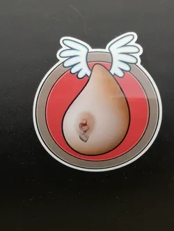 The sticker features a stylized depiction of a human breast with a nipple piercing. The breast is rendered in realistic skin tones (light beige/peach) and is adorned with a small silver-colored ring piercing from which a tiny silver heart charm dangles. Above the breast, two cartoon-style white wings with light blue outlines extend outwards. The central image is framed by a series of concentric circles: an inner red circle, followed by a thin black line, a wider grey band, and finally a white outer border. The overall design is playful and suggestive, combining anatomical realism with whimsical elements.